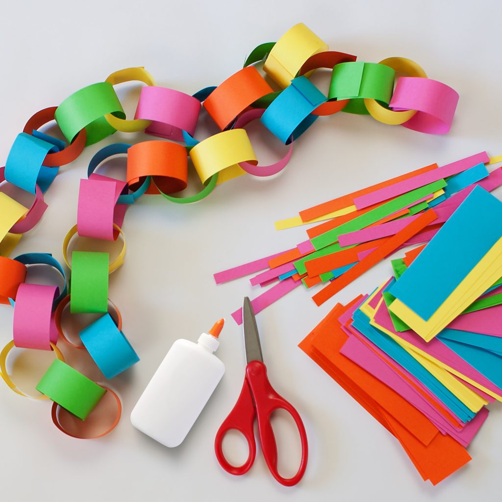 Colorful paper chain made with red, green, blue, pink, orange, and yellow paper strips, shown alongside glue, scissors, and additional strips of paper on a crafting table.