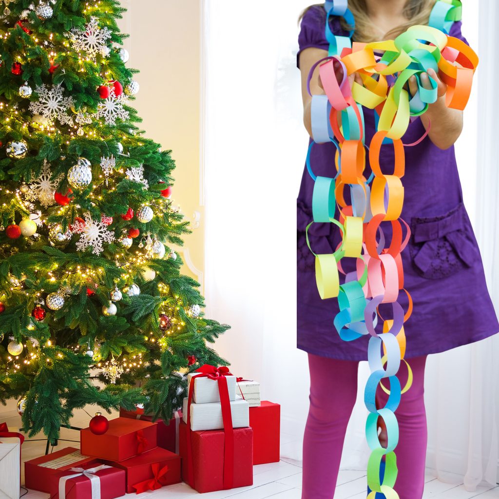 A child holding a colorful paper chain in front of a decorated Christmas tree with ornaments, lights, and presents beneath it.